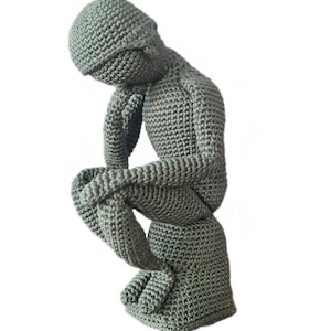 May include: A handmade, crocheted sculpture of Rodin's The Thinker in a muted green color. The figure is seated, with its chin resting on its hand, and is made with a textured stitch. The sculpture is a unique piece of home decor.