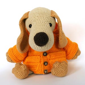Può includere: Un cane di peluche lavorato a uncinetto che indossa una giacca arancione brillante con bottoni. Il cane ha gli occhi marroni e un naso marrone.