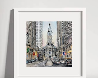 May include: Watercolor painting of a city street scene, framed in white. The artwork depicts a grand building with a clock tower, flanked by tall buildings. Cars line the street, and colorful flags hang from the buildings.