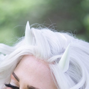 以下が含まれることがあります： 白い髪と白い角を持つ女性が、透き通るメッシュのオーバーレイが付いた黒いトップを着ています。トップには「The Shimmering Chimera」という文字が見えます。