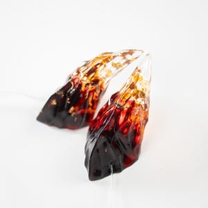 Puede incluir: Dos esculturas abstractas de resina con forma de llama. Las esculturas transitan de un rojo oscuro intenso en la base a un naranja y dorado vibrantes en la parte superior. Las piezas se colocan sobre un fondo blanco limpio, resaltando sus colores vibrantes y su forma única.
