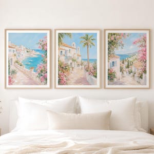 May include: A set of three framed wall art prints featuring oil-style paintings of Mediterranean coastal scenes with white buildings, blue water, palm trees, and vibrant pink bougainvillea flowers, displayed in light wood frames on a wall.