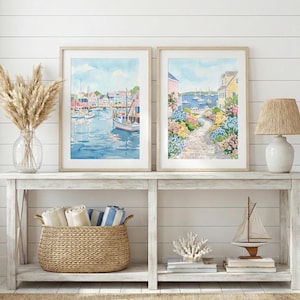 May include: Two framed watercolor paintings depicting coastal scenes. One shows a harbor with boats and buildings, the other a path lined with flowers leading to the sea. Both are in light wood frames, displayed on a white console table.