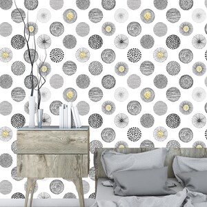 May include: A bedroom with a white wall featuring a black and gold geometric pattern wallpaper. The wallpaper has a repeating pattern of circles with different textures and designs. A wooden nightstand with two drawers sits in front of the wall. A gray bed with a gray duvet and two pillows is in the foreground.