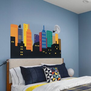 May include: A blue bedroom with a bed and a cityscape wall decal. The decal features a black silhouette of a city with colourful buildings and a helicopter flying over a full moon.