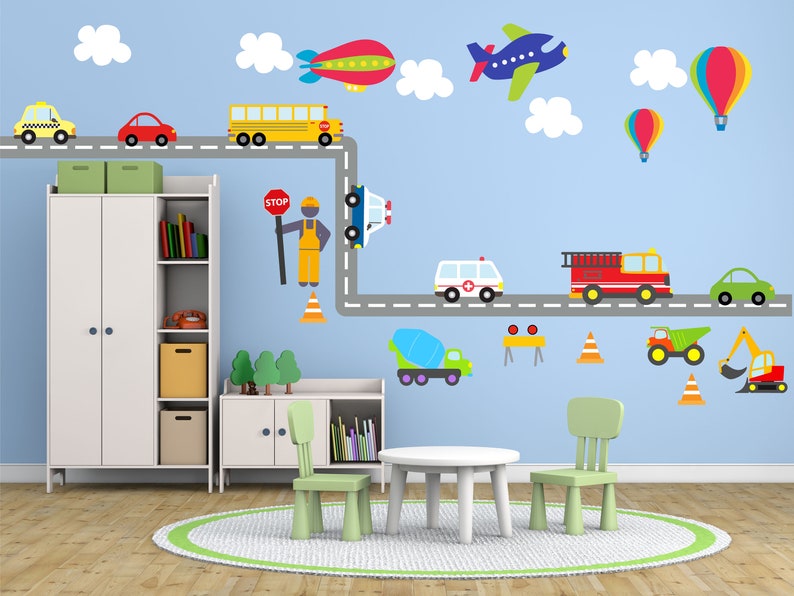 May include: A blue wall with a road and various vehicles, including a school bus, ambulance, fire truck, and construction vehicles. There are also clouds, a hot air balloon, and an airplane. The wall is decorated with a road and traffic signs. There are two green chairs and a white table in front of the wall.