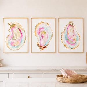 May include: Three framed wall art prints featuring watercolor illustrations of oyster shells. Each shell displays a vibrant palette of pink, blue, and gold tones against a white background, enclosed in thin gold frames hanging on a wall.