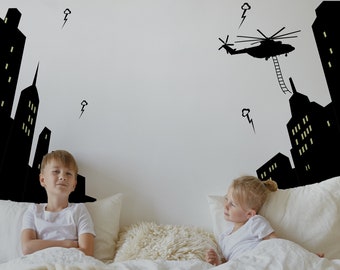 Op de afbeelding: Twee kinderen liggen in bed en kijken naar een muur met zwarte en witte stickers van een skyline en een helikopter. De stickers zijn glow-in-the-dark.