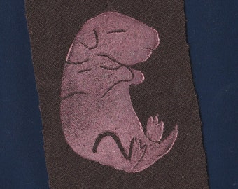 May include: A rectangular dark brown fabric patch featuring a centered, stylized illustration of a small, curled-up creature in a muted mauve color. The creature is depicted in a fetal position with visible outlines of its head, limbs, and tail.