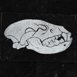 Pode incluir: Um remendo bordado branco em um fundo de tecido preto representando uma caveira detalhada de um animal com dentes afiados.