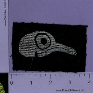 May include: A black fabric patch with a white embroidered bird head design. The bird has a large eye and a long beak.
