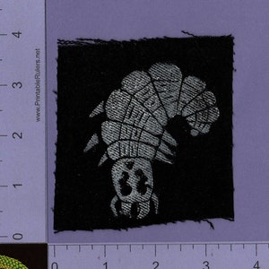 May include: A black fabric patch with a white embroidered image of a caterpillar. The caterpillar is facing left and has a segmented body.