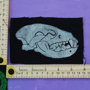 May include: A rectangular black fabric patch featuring a white embroidered animal skull in profile. The patch is positioned against two wooden rulers, showing it measures approximately 4.5 inches in width and 2.5 inches in height.