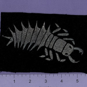 May include: A black fabric patch with a silver embroidered design of a stylized creature with a segmented body and a sharp, pointed head.