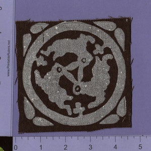 May include: A square patch of fabric with a silver design on a brown background. The design is a circle with a stylized floral pattern inside. The patch is approximately 2 inches by 2 inches.
