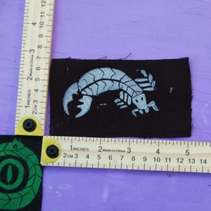 May include: A rectangular black fabric patch featuring a silver-colored scorpion graphic. The patch is positioned against a wooden ruler showing a width of approximately 3.5 inches and a height of approximately 2 inches for the main design.