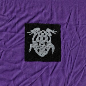 May include: A black fabric patch with a white embroidered image of a frog. The frog is facing up with its legs spread out.