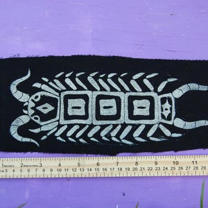 May include: A rectangular black fabric patch featuring a white screen-printed graphic of a segmented, multi-legged creature with horns. A yellow ruler shows the patch is approximately 8.5 inches long and 3 inches wide at its widest point.