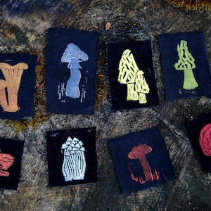 May include: A collection of eight small, square fabric patches with various mushroom designs in metallic gold, silver, and red colors. The patches are set against a dark background, with the image taken on a tree stump.