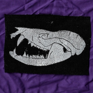 May include: A black rectangular patch featuring a stylized, white illustration of a skull with visible teeth. The skull is depicted in profile, with the mouth open. The patch is likely made of fabric and is set against a purple background.