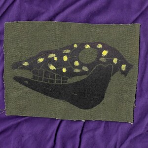 May include: A green fabric patch with a black silhouette of an animal skull. The skull is decorated with yellow paint splatters.