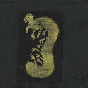 May include: A black fabric patch with a yellow silhouette of a creature with multiple limbs and a long tail.