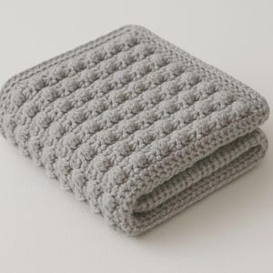 May include: Folded gray crocheted blanket with a textured, bobble stitch pattern. The blanket is made of soft yarn and has a rectangular shape. The edges are neatly finished, and the blanket appears to be folded in half.