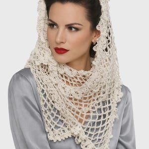 May include: An off-white triangular crochet shawl featuring an intricate openwork mesh pattern and a decorative scalloped border. The lightweight textile is draped over the head and shoulders, showcasing a detailed floral-inspired lace edge.