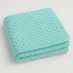 Può includere: Una coperta quadrata all'uncinetto di colore turchese piegata. La coperta ha un motivo strutturato ed è realizzata in filato morbido. I bordi sono rifiniti con cura e la coperta è piegata in tre parti, mostrando il suo spessore e la sua costruzione. La coperta è adatta a un bambino.