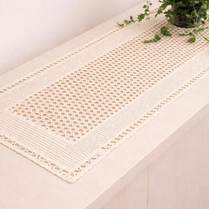 Op de afbeelding: Een crèmekleurige, rechthoekige tafelloper met een delicaat, open geweven ontwerp. De loper heeft een patroonrand en is geplaatst op een lichtgekleurd houten oppervlak. Bovenop staat een kleine plant in een glazen vaas.