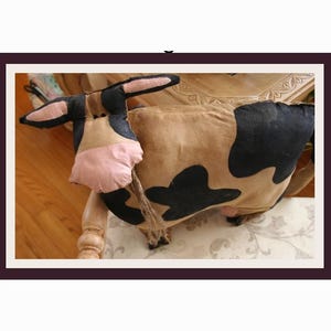 May include: A handmade cow-shaped decorative pillow. The pillow features a tan body with black spots, a pink snout, and black ears. A rope tail hangs from the back. The pillow is resting on a chair with a floral patterned seat.