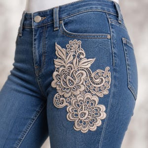 Peut inclure: Jean bleu avec une application de dentelle florale beige sur la poche avant. L'application présente des motifs floraux et de feuilles détaillés. Le jean a une conception classique à cinq poches et une fermeture à bouton et fermeture éclair.