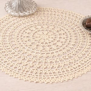 May include: A cream-colored, circular, crocheted doily with an intricate floral pattern. A silver-colored dish with chocolate candies sits on the right. A decorative glass object is in the upper left.