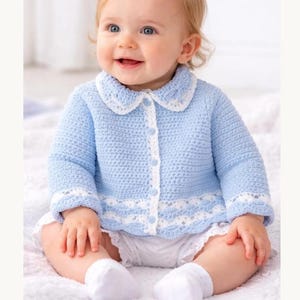 May include: A baby in a light blue crochet cardigan with white detailing, a matching collar, white shorts and socks. The cardigan is buttoned. The baby is smiling.