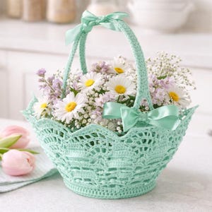 May include: A light mint green crocheted basket with a scalloped rim and a single handle. The basket is decorated with two matching mint green satin ribbons tied at the base of the handle and at the top, filled with white daisies and purple flowers.