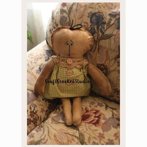 May include: A handmade fabric doll with a tan head and limbs, wearing a green gingham dress with a lace collar. The doll has a simple face with stitched features and a small black bow. The text "Craft Crochet Studio" is on the dress.
