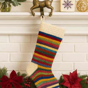 May include: A vibrant, crocheted Christmas stocking with a cream-coloured cuff and toe, featuring a striped pattern in multiple colours. The stocking is suspended from a bronze reindeer hook on a white brick wall, adorned with festive greenery and a red bow.