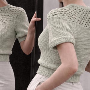 Puede incluir: Un top de ganchillo verde claro con mangas cortas y un diseño texturizado. El top presenta un cuello redondo y una cintura ajustada. La parte superior del pecho tiene un patrón de tejido abierto único. El top se combina con pantalones de color claro.