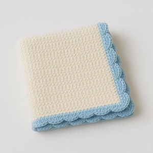 Può includere: Una coperta di crochet piegata, color crema, con un bordo smerlato azzurro. La coperta è quadrata e sembra morbida e accogliente, adatta a un bambino. Il punto crochet crea una superficie strutturata.