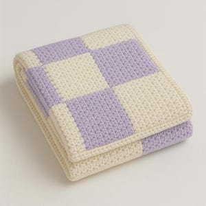 Può includere: Coperta in crochet piegata con motivo a scacchiera in lavanda e crema. La coperta è realizzata con un filato spesso e strutturato. I bordi piegati mostrano la qualità artigianale. Un elemento d'arredo accogliente.