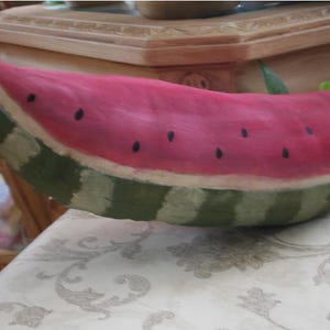 May include: A watermelon-shaped decorative item with a pink interior and green striped exterior. The watermelon has black seed details and is resting on a patterned surface. The item is likely a decorative piece for a kitchen or dining area.