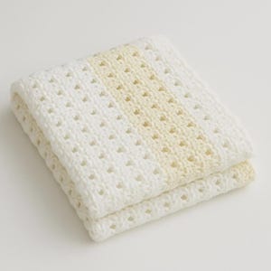 May include: Folded white knitted blanket with a central stripe of pale yellow. The blanket has a textured, open weave design, and is folded neatly. The soft color palette and texture suggest a cozy, handmade item.