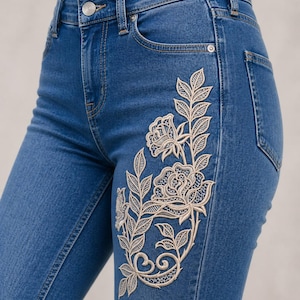 Op de afbeelding: Blauwe jeans met crèmekleurige bloemenborduurwerk aan de voorkant. De borduursels tonen een rozen- en bladerenpatroon. De jeans hebben een klassiek ontwerp met vijf zakken en een knoop- en ritssluiting.