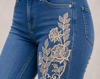 May include: Blue denim jeans with an embroidered floral design on the front. The embroidery features a cream-colored rose and leaf pattern. The jeans have a classic five-pocket design and a button and zipper closure.