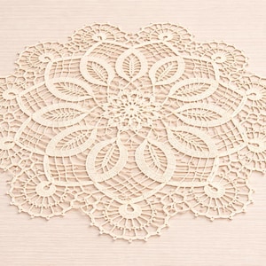 Può includere: Tovagliolo circolare color crema con un intricato design traforato. Il tovagliolo presenta un motivo floreale centrale circondato da motivi ripetuti a forma di foglia e delicati bordi smerlati. Il tessuto fatto a mano è posto su una superficie di legno chiaro.