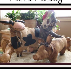 Op de afbeelding: Handgemaakte boerderijdierensculpturen, waaronder een koe met zwarte vlekken, een varken en een haan. De koe heeft een roze neus en oren. De sculpturen worden tentoongesteld op een oppervlak met patronen.