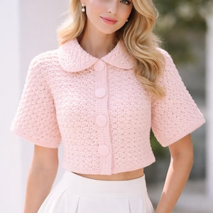 May include: A pale pink, short-sleeved, crocheted cardigan with a rounded collar and large buttons. The cropped top is paired with a white pleated skirt. The cardigan is a soft, pastel colour.