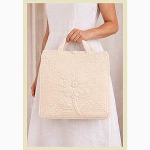 Pode incluir: Bolsa tote quadrada creme com um design floral de crochê. A bolsa apresenta um motivo floral central com pétalas e folhas detalhadas. Possui duas alças e uma textura tecida, sugerindo uma qualidade artesanal.