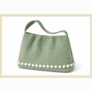 May include: A sage green crochet handbag with a single handle. The bag is rectangular with a row of white pom-poms along the bottom edge. The bag is made of a soft, textured yarn.