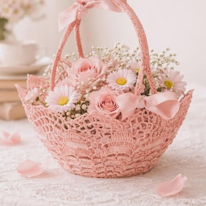 Könnte beinhalten: Ein hellrosa gehäkelter Korb mit passendem Henkel und einer Schleife aus Satinband. Der Korb ist mit einem Blumenarrangement gefüllt, das aus hellrosa Rosen, weißen Gänseblümchen mit gelber Mitte und kleinen weißen Schleierkrautblüten besteht.